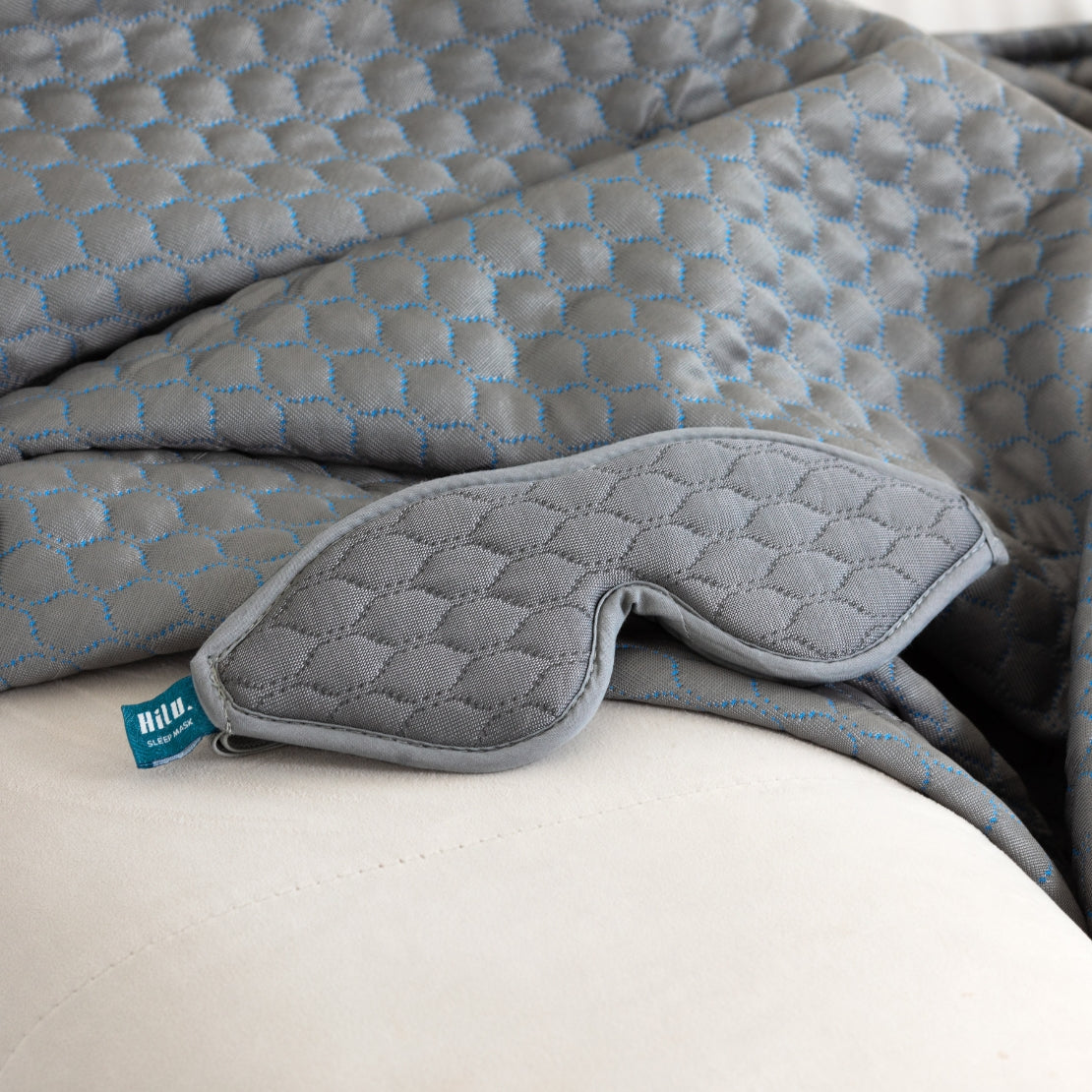 A close-up view of a gray HILU sleeping mask with a quilted pattern, placed atop a matching quilted blanket. The mask features the 'Hilu' brand label in blue.
