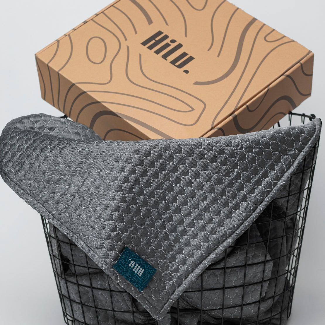 A gray quilted blanket with the brand label 'HILU' draped over a black metal basket. Above it, a uniquely designed cardboard box with 'HILU' printed on it, showcasing abstract wavy patterns.